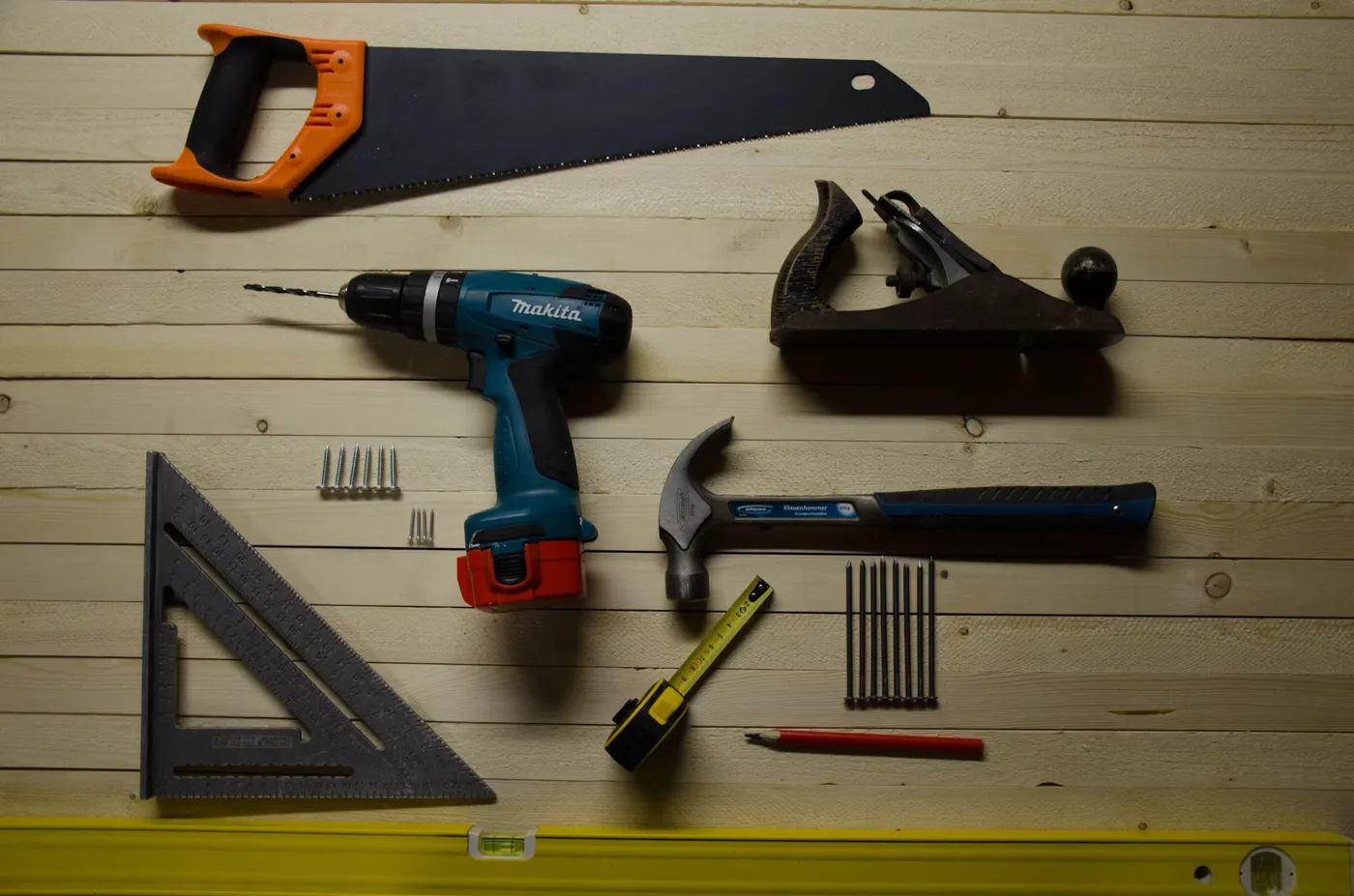 Akku-Bohrschrauber, Handsäge, Hobel, Hammer, Maßband und Winkel auf einer Holzwand